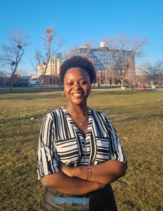 A woman stands in front of a park smiling at the camera with her arms folded. 