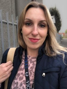 A white woman with blonde hair smiles into the camera - this is a selfie. She wears a dark blue jacket and a VICTVS lanyard with a cream handbag.